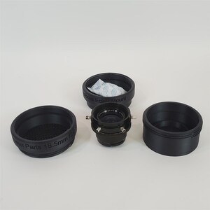 May include: A black camera lens with a 18.5mm focal length, a black focus ring, and a black lens cap. The lens is labeled "Focus Eclair Mount" and "Aux Paris 18.5mm f2".