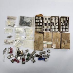 May include: A collection of electronic quartz crystal oscillators, including several small plastic bags with handwritten labels, four cardboard storage boxes with grid inserts, and loose components in various metallic and colored finishes.