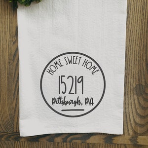 May include: White kitchen towel with the text "HOME SWEET HOME" and the number "15219" inside a black circle. The text "Pittsburgh, PA" is below the number. A green wreath is in the upper left corner.