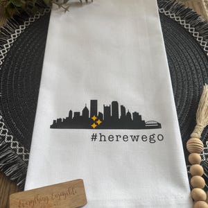 May include: White kitchen towel with a black skyline graphic, two yellow stars, and the text "#herewego". A small wooden sign with the text "Everything Enjoyable Designs" is also present.