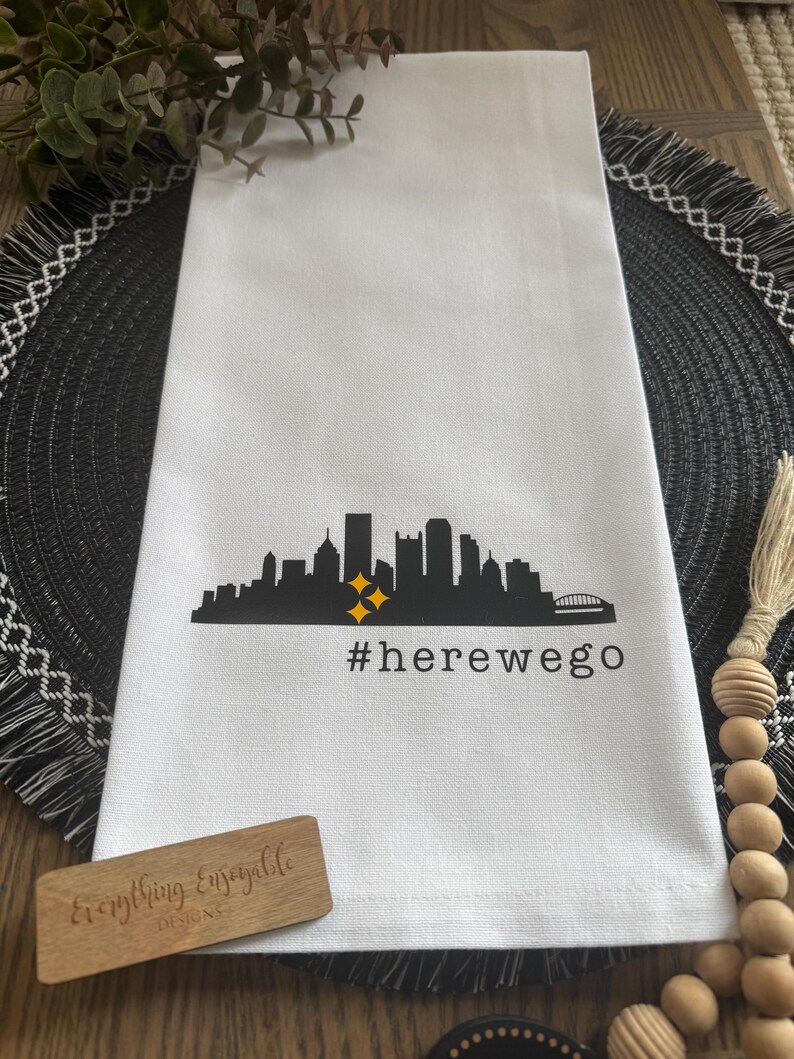 May include: White kitchen towel with a black skyline graphic, two yellow stars, and the text "#herewego". A small wooden sign with the text "Everything Enjoyable Designs" is also present.