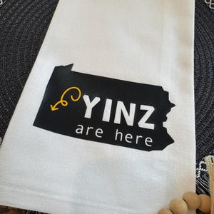 May include: White kitchen towel with the text "YINZ are here" in black and white, with a yellow arrow and a black outline of Pennsylvania. The towel is folded and placed on a dark gray placemat.