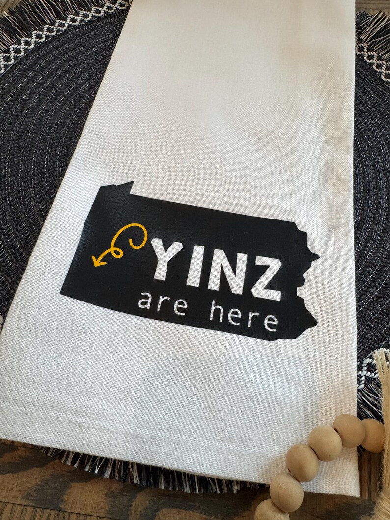 May include: White kitchen towel with the text "YINZ are here" in black and white, with a yellow arrow and a black outline of Pennsylvania. The towel is folded and placed on a dark gray placemat.