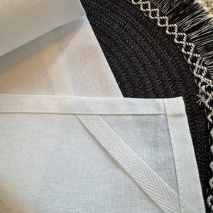 May include: Close-up of a white linen kitchen towel with a stitched border and a hanging loop. The towel is folded, revealing the texture of the fabric. A black woven placemat with a white geometric border is in the background.