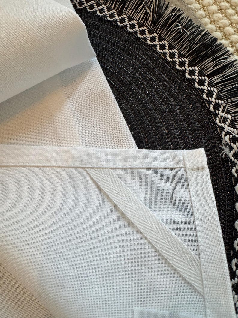 May include: Close-up of a white linen kitchen towel with a stitched border and a hanging loop. The towel is folded, revealing the texture of the fabric. A black woven placemat with a white geometric border is in the background.