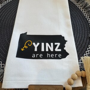 May include: White kitchen towel with a black Pennsylvania outline and the words "YINZ are here" in white. A yellow arrow points to the text. The towel is on a black and gray placemat.