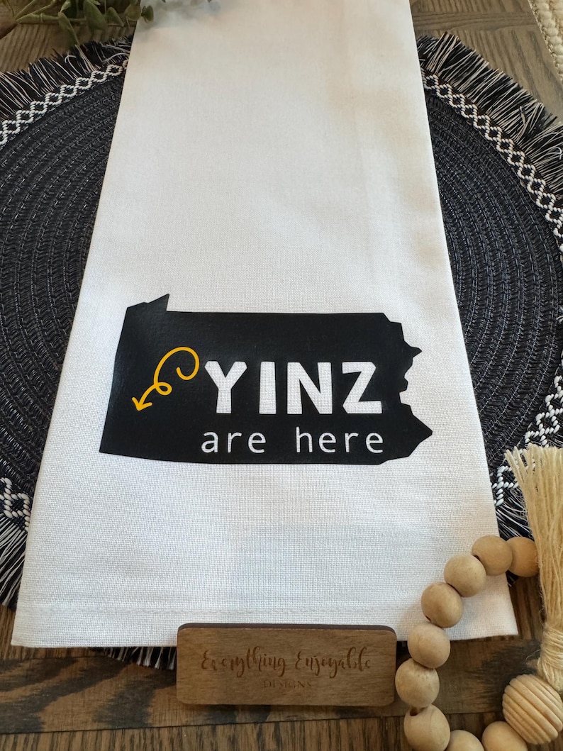 May include: White kitchen towel with a black Pennsylvania outline and the words "YINZ are here" in white. A yellow arrow points to the text. The towel is on a black and gray placemat.