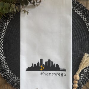 May include: White kitchen towel featuring a black silhouette of a city skyline with two yellow stars. The text "#herewego" is printed below the skyline. The towel is made of a soft, absorbent material.