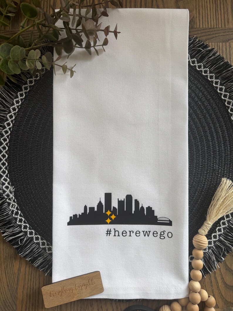 May include: White kitchen towel featuring a black silhouette of a city skyline with two yellow stars. The text "#herewego" is printed below the skyline. The towel is made of a soft, absorbent material.