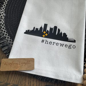 May include: White kitchen towel with a black skyline graphic, two yellow starbursts, and the text "#herewego". A small wooden sign with the text "Everything Enseayable Designs" is also visible.