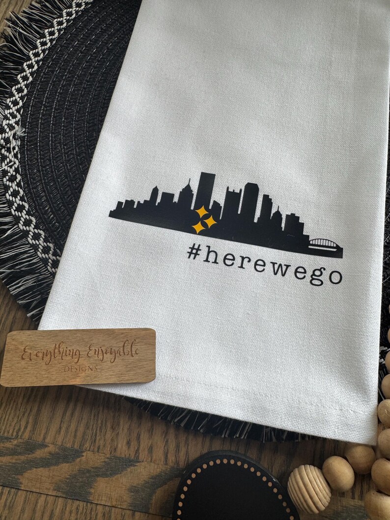 May include: White kitchen towel with a black skyline graphic, two yellow starbursts, and the text "#herewego". A small wooden sign with the text "Everything Enseayable Designs" is also visible.