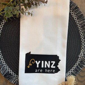 May include: White kitchen towel with a black and yellow graphic of the state of Pennsylvania and the words "YINZ are here". The towel is folded and placed on a black placemat with a decorative fringe.
