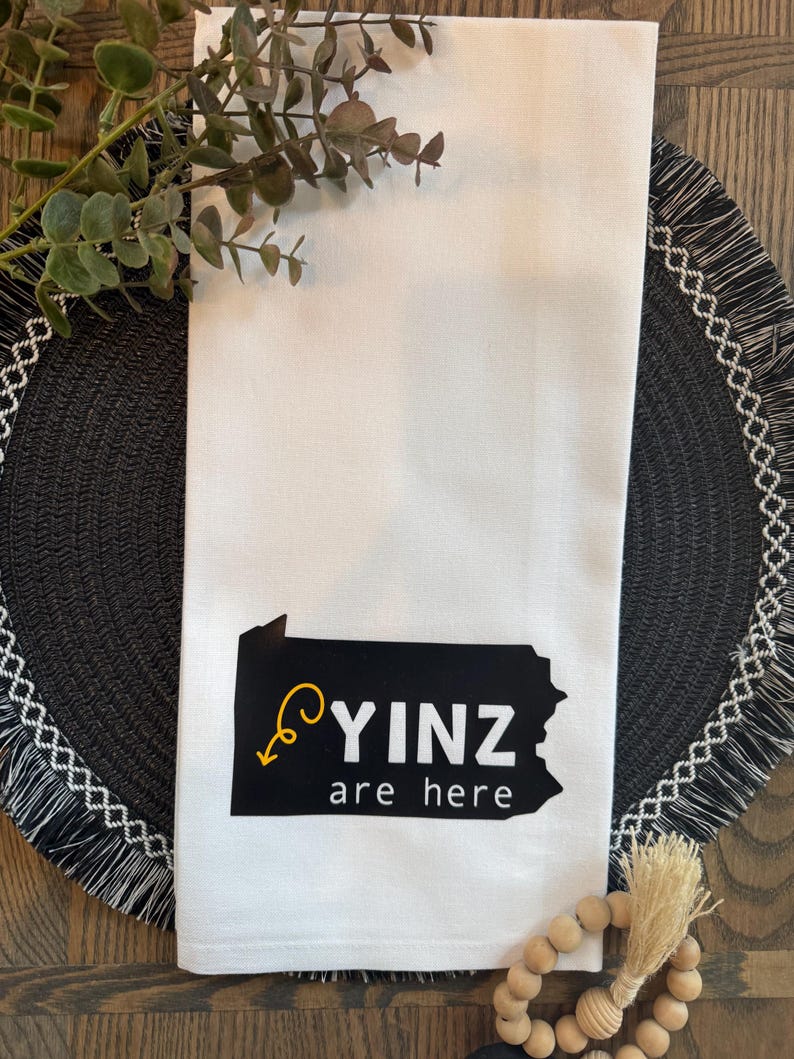 May include: White kitchen towel with a black and yellow graphic of the state of Pennsylvania and the words "YINZ are here". The towel is folded and placed on a black placemat with a decorative fringe.