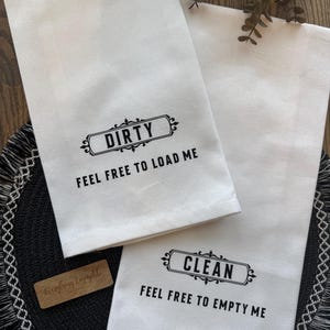 May include: Two white kitchen towels with black text. One towel reads "DIRTY FEEL FREE TO LOAD ME" and the other reads "CLEAN FEEL FREE TO EMPTY ME". A small wooden tag is also visible.