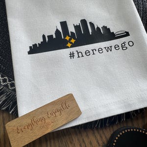 May include: White kitchen towel with a black skyline graphic, two yellow stars, and the text "#herewego". A wooden sign with the text "Everything Enjoyable DESIGNS" is also visible.