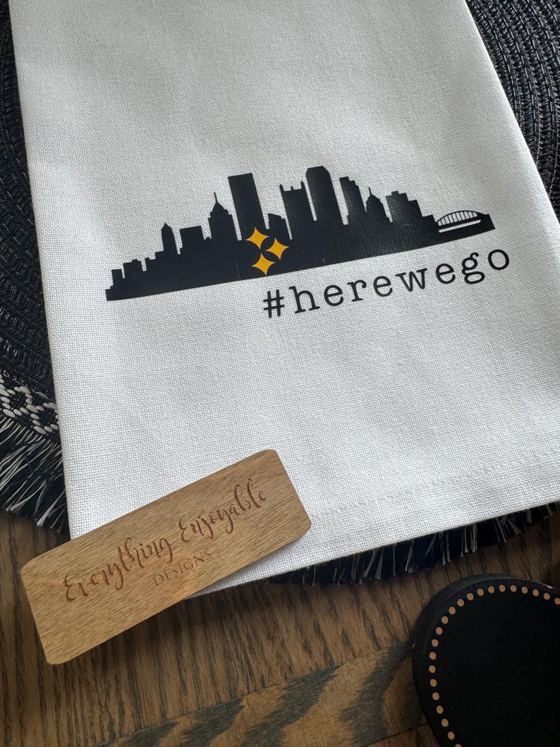May include: White kitchen towel with a black skyline graphic, two yellow stars, and the text "#herewego". A wooden sign with the text "Everything Enjoyable DESIGNS" is also visible.