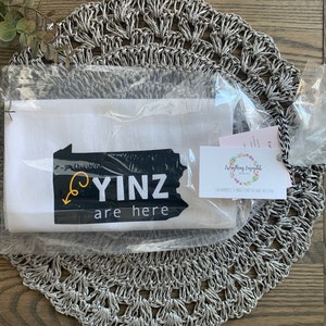 May include: A white tea towel with the text "YINZ are here" in black and yellow, packaged in clear plastic. The towel features a graphic of the state of Pennsylvania. A small tag with floral design is attached.