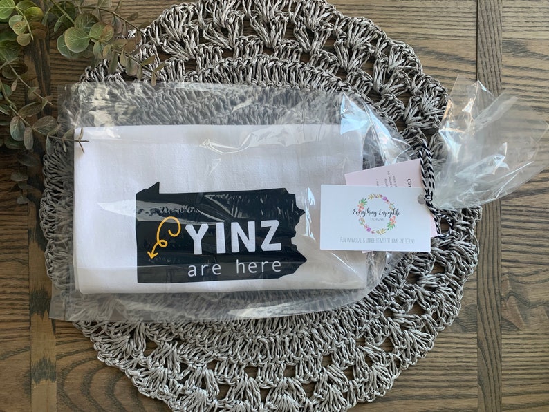 May include: A white tea towel with the text "YINZ are here" in black and yellow, packaged in clear plastic. The towel features a graphic of the state of Pennsylvania. A small tag with floral design is attached.