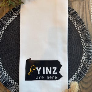 May include: White kitchen towel with a black graphic of the state of Pennsylvania and the words "YINZ are here" in black and yellow. The towel is folded and placed on a decorative table setting.