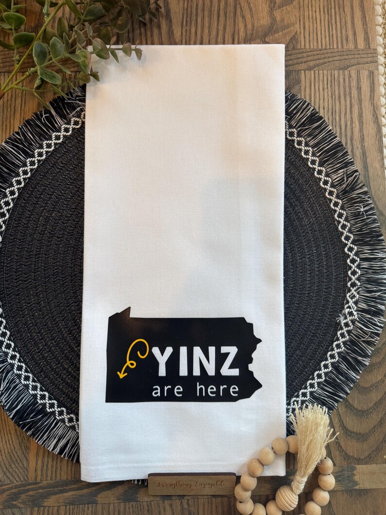 May include: White kitchen towel with a black graphic of the state of Pennsylvania and the words "YINZ are here" in black and yellow. The towel is folded and placed on a decorative table setting.