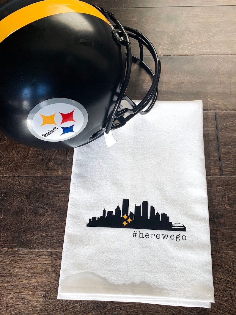 May include: A black and gold football helmet with the Steelers logo rests next to a white kitchen towel. The towel features a black silhouette of a city skyline with two gold stars and the hashtag #herewego.