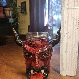 Puede incluir: Una taza de cerámica roja con forma de diablo con cuernos dorados y una lengua roja larga. La taza mide aproximadamente 15 cm de alto.