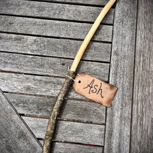 May include: A light brown wooden stick with a rough texture, labeled "Ash" on a brown tag.