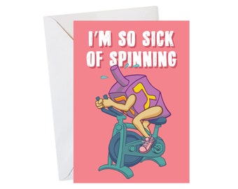 May include: A pink greeting card with a cartoon illustration of a dreidel riding a stationary bike. The text on the card reads "I'm so sick of spinning."
