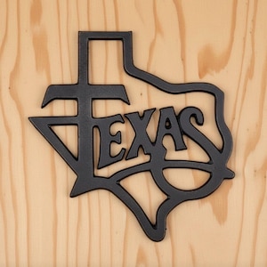 May include: A black metal wall art piece shaped like the outline of the state of Texas. The design features the word TEXAS integrated into the centre of the state shape in a bold, stylised font, with the letter T forming a cross-like structure.