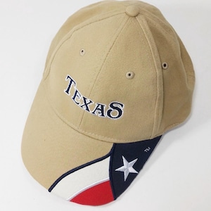 Może przedstawiać: Beżowa czapka z daszkiem z napisem Texas wyhaftowanym ciemnoniebieskimi literami z przodu. Daszek posiada ukośny wzór z białymi i czerwonymi pasami, ciemnoniebieską sekcją oraz wyhaftowaną białą gwiazdą z małym symbolem TM.