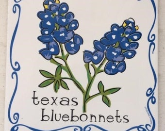 May include: A white ceramic tile with a blue and green illustration of Texas bluebonnets. The tile has a blue border with a decorative scroll design. The text "texas bluebonnets" is written in black below the flowers.