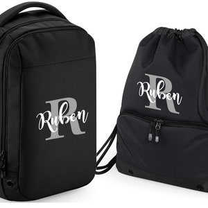May include: Black backpack and drawstring bag set with the name "Ruben" and a large "R" monogrammed in white on each bag.