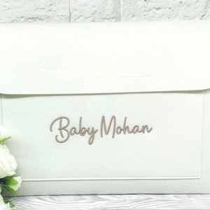 May include: A cream-coloured baby clutch bag with a flap closure. The text "Baby Mohan" is embroidered in a cursive font on the front. The bag is rectangular with a stitched border. White roses are in the lower left corner.