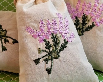 May include: Three white cotton drawstring bags with embroidered purple and green flowers. The bags are on a green patterned surface.