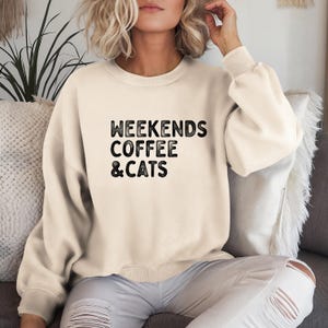 Könnte beinhalten: Ein cremefarbener Pullover mit dem Aufdruck "Weekends Coffee & Cats" in Schwarz.