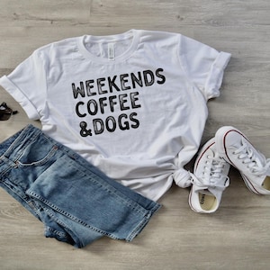 May include: A white t-shirt with the text "WEEKENDS COFFEE & DOGS" in black, paired with blue jeans, white sneakers, and sunglasses. The casual outfit is laid out on a light wood-grain surface.