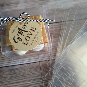 May include: A clear plastic box filled with a s'more treat. The s'more is made with a graham cracker, marshmallow, and chocolate. The box has a tag attached to it that says "S'more Love" with the names "Tiger & McKenna" and the date "9.21.24". The tag also says "Take Home".