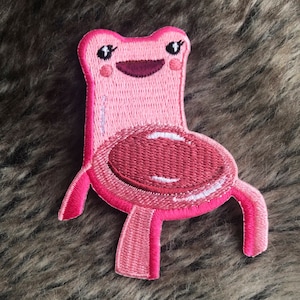 Könnte beinhalten: Rosa gestickter Aufnäher einer Frosch-förmigen Stuhl mit einem lächelnden Gesicht und großen Augen.