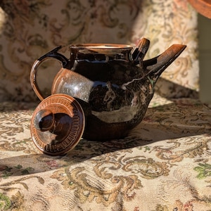 May include: A dark brown ceramic teapot with a lid, handcrafted. The teapot has a glossy finish and a unique spout design. The lid is off to the side, revealing the interior. The teapot is on a floral patterned surface.