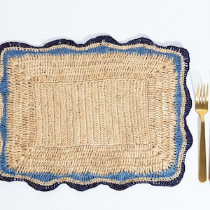 May include: A woven placemat with a blue and black border. The placemat has a scalloped edge and is made of natural fibers. It is shown with a gold fork and knife.