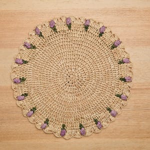 May include: A round, beige crochet doily with a scalloped edge. The edge is adorned with small, purple flower buds with green stems. The doily is placed on a light brown wooden surface.