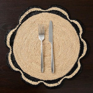 May include: A round, beige placemat with a black crocheted border. The placemat has a scalloped edge and is made of a woven material. A silver fork and knife are placed on the placemat.