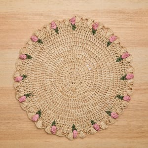 May include: A round, beige crocheted doily with a scalloped edge. Pink and green floral accents are evenly spaced around the border. The doily is made of a natural fiber and sits on a light wood surface.