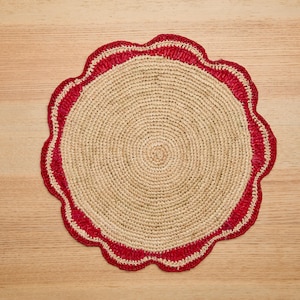May include: A round, crocheted placemat with a scalloped edge. The placemat is made of natural fibers and has a red border.