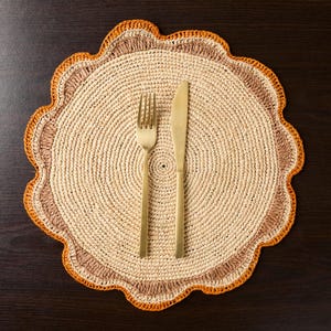 May include: A round, crocheted placemat with a brown and orange border. A gold fork and knife are placed on the placemat.