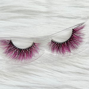 Könnte beinhalten: Ein Paar synthetische Wimpern mit schwarzer Basis und einem Farbverlauf aus rosa und schwarzen Fasern. Die Wimpern werden in einer transparenten, dreieckigen Kunststoffhülle vor einem flauschigen, weißen Hintergrund präsentiert.