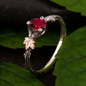 Könnte beinhalten: Ein silberner Ring mit einem roten Edelstein und einem goldenen Blattdesign. Der Ring liegt auf einem Bett aus grünen Blättern.