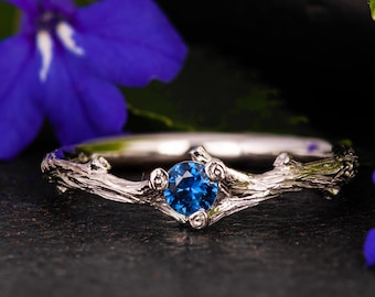 May include: A silver ring with a blue gemstone set in a branch-like design. The ring is surrounded by blue flowers and green leaves.