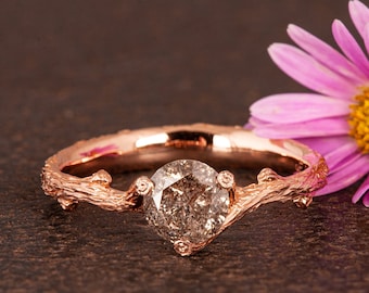 May include: A rose gold engagement ring featuring a brown diamond set in a twig-like band. The ring is resting on a brown surface beside a pink flower.