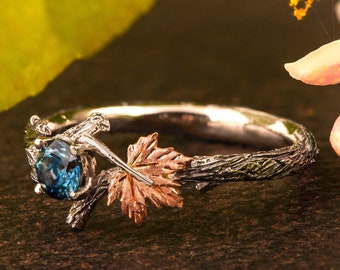 May include: A silver ring with a blue gemstone set in a twig design. The ring features a rose gold leaf on one side.
